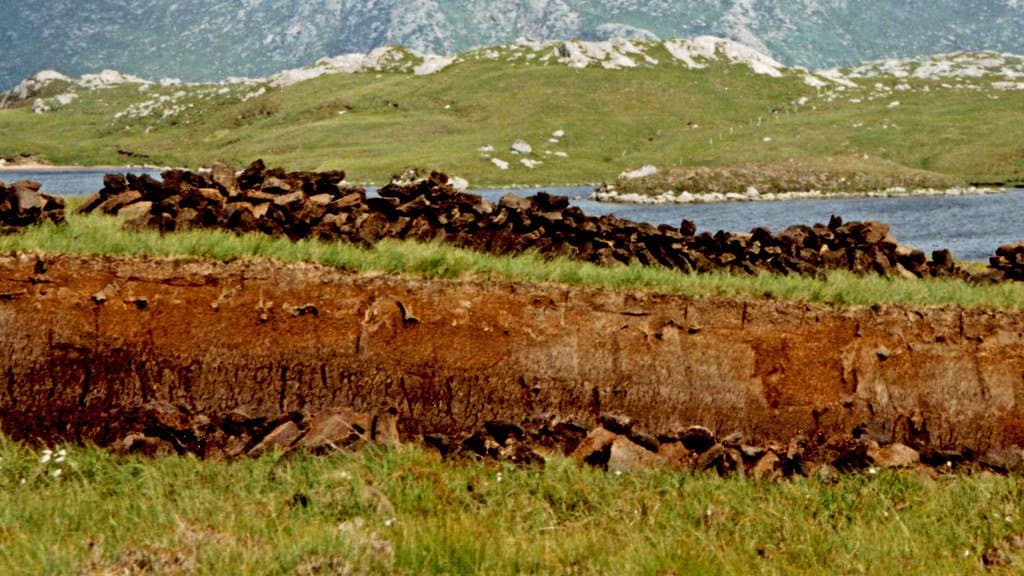 An exposed face of cut peat about a metre deep runs across a moorland foreground, with stacked drying peat turves along its top, a loch behind it and hills beyond.