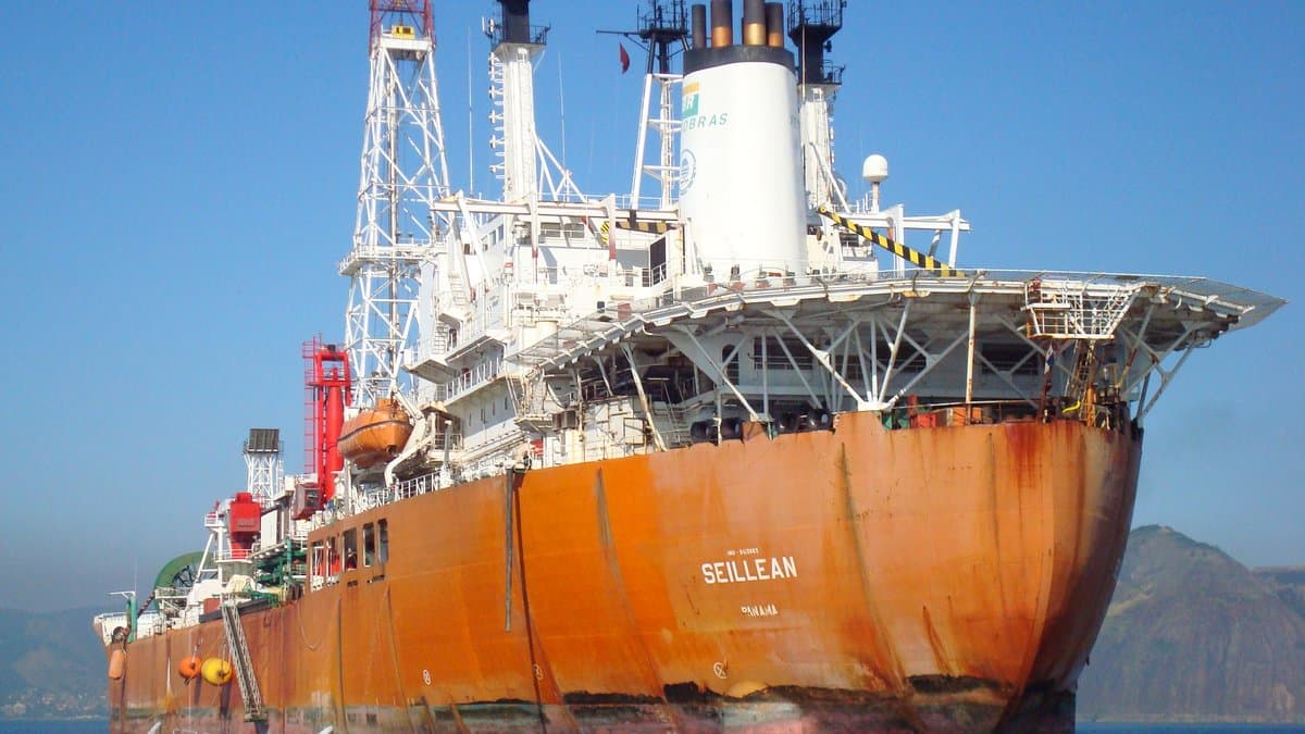 A large rust-orange floating production, storage and offloading vessel named Seillean lies at anchor in calm blue water off a Brazilian coastline, its deck crowded with cranes, a derrick and pipework.