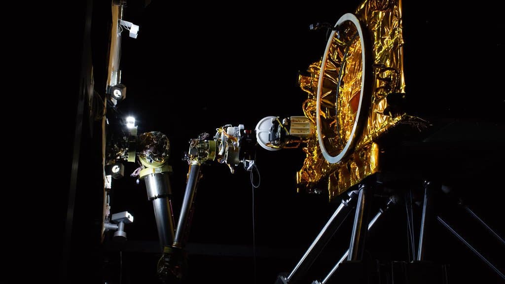 A robotic arm extends toward a gold-foil-wrapped satellite mock-up in a darkened spacecraft servicing test facility.