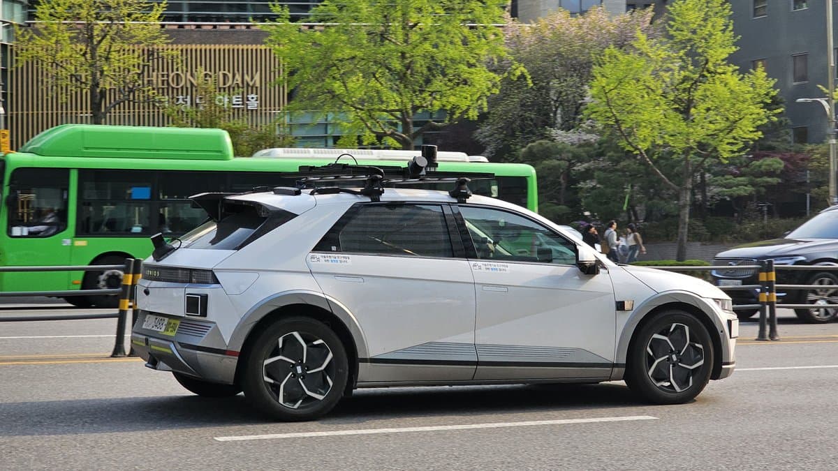 A white Hyundai IONIQ 5 robotaxi with a roof-mounted sensor pod of lidar units and cameras drives along a city street past a green bus.