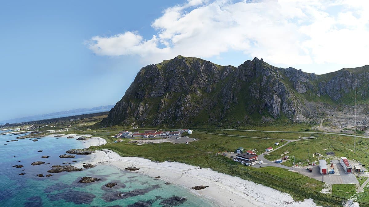 Aerial panorama of Andøya Space in northern Norway: low buildings and launch facilities on a green coastal plain between a white-sand beach and a steep mountain ridge.