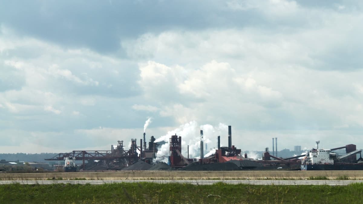 An integrated steel mill on a waterfront, with blast-furnace structures, tall stacks venting white plumes, ore stockpiles and a bulk carrier moored alongside.