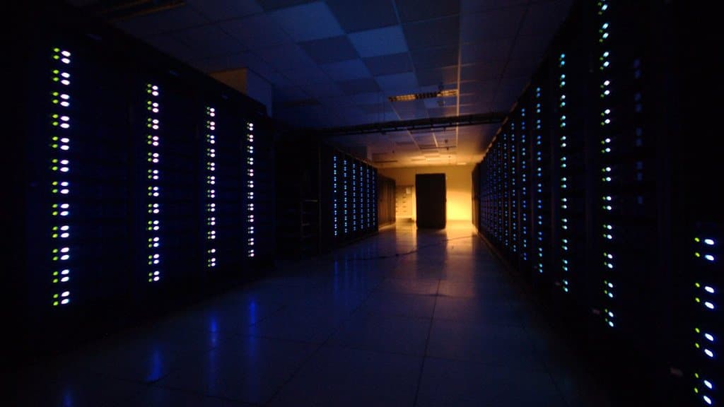 A dark data-centre aisle lined floor to ceiling with computer racks, their status lights glowing in rows on both sides