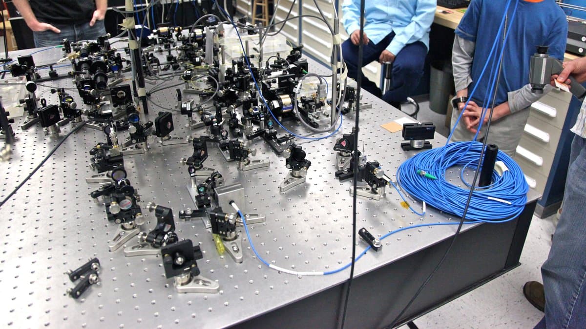 A laser optical table in a physics laboratory crowded with mirrors, lenses and precision mounts, with a large coil of blue fibre-optic cable at the edge of the bench.
