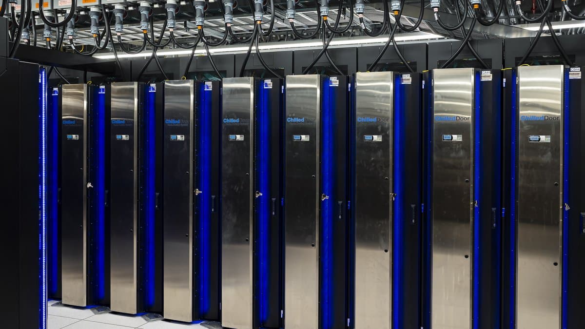Rows of tall black computer cabinets with polished metal cooling doors and blue indicator strips in a supercomputer machine hall, overhead power connectors above them