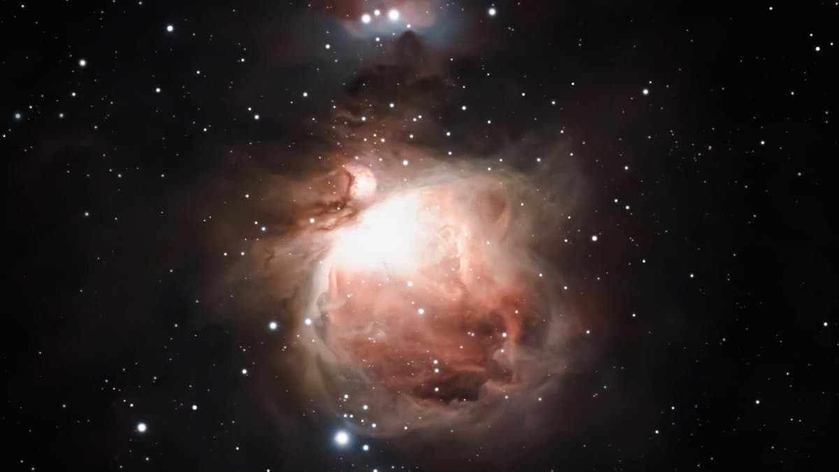 Deep telescopic photograph of the Orion Nebula: glowing pink and white clouds of gas and dust around a bright core, with scattered stars on a black sky.