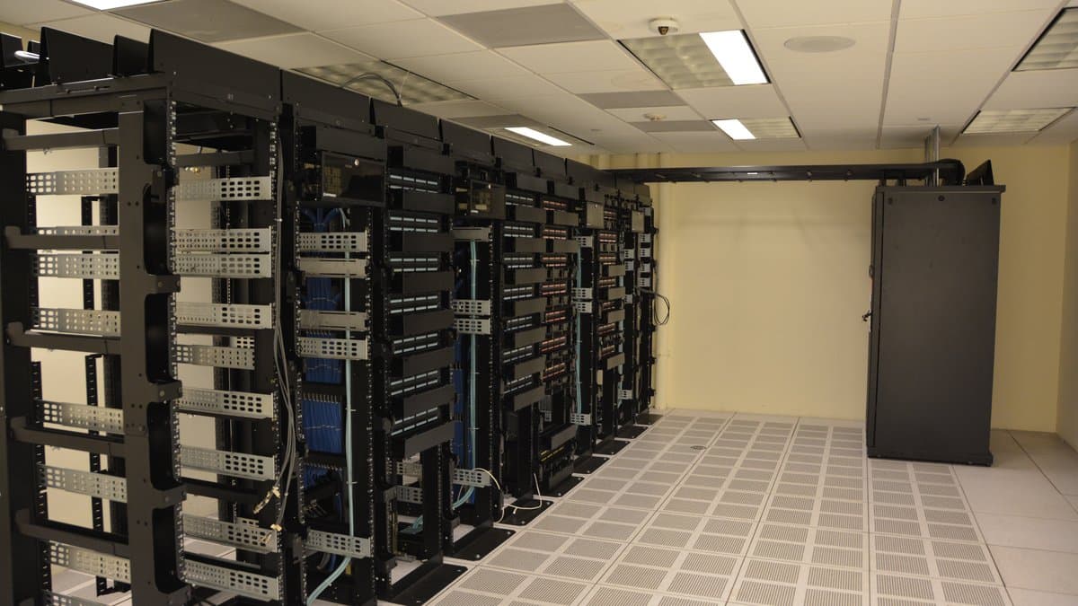 Rows of black server racks with network cabling in a small data centre, raised floor tiles in the foreground