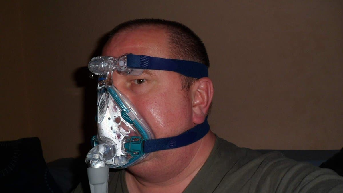 A man reclining with a blue-strapped full-face CPAP mask over his nose and mouth, a ribbed air hose leading away from it.