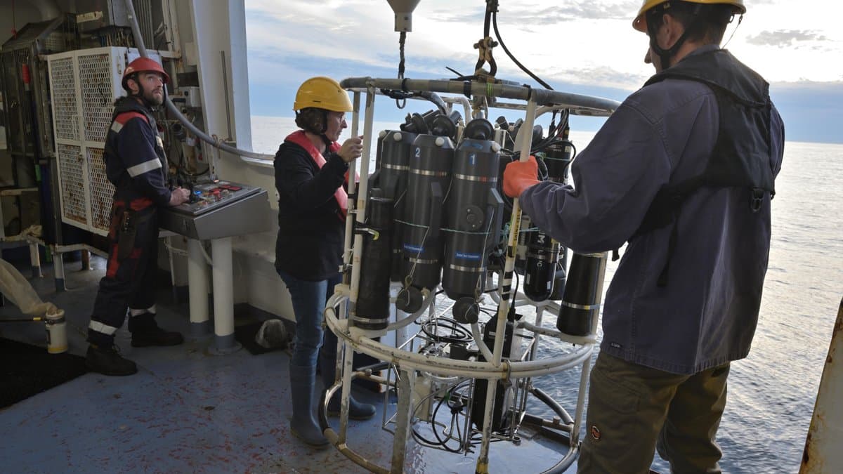Three crew in hard hats on a research ship's deck handle a CTD rosette of grey water-sampling bottles suspended over calm open sea.