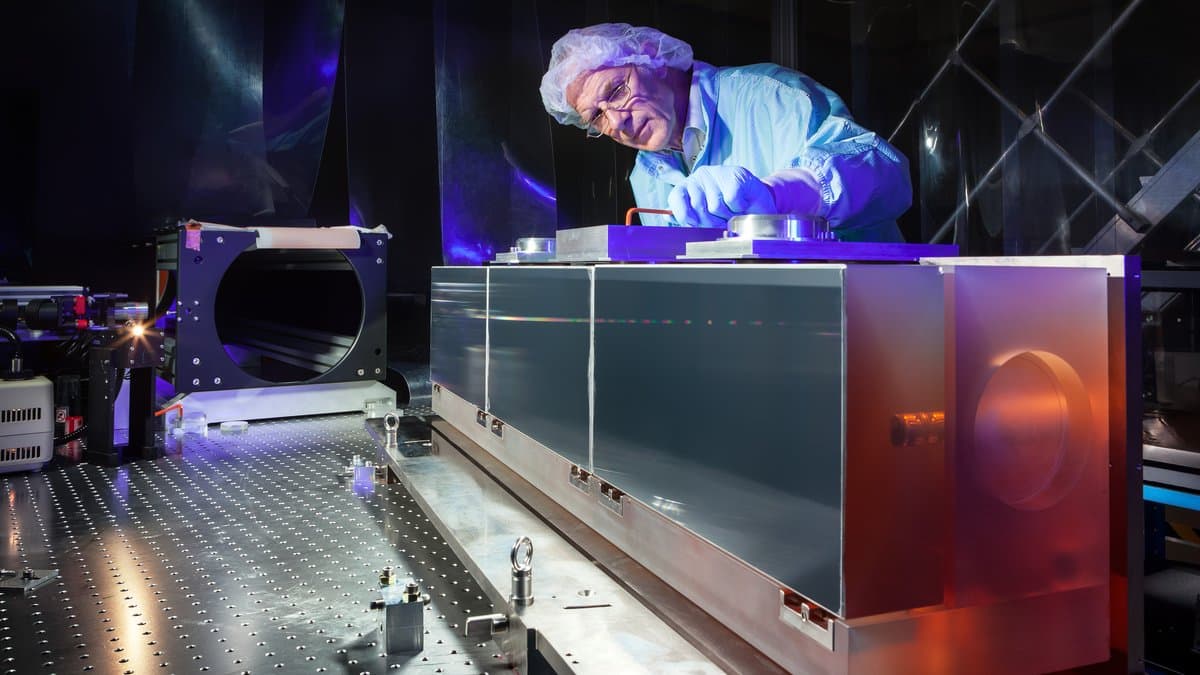 An engineer in a cleanroom suit and hairnet leans over the long metal housing of the ESPRESSO spectrograph's diffraction grating in a darkened laboratory illuminated in blue and violet light.