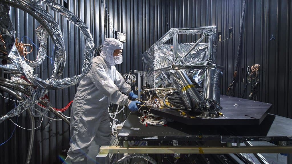 An engineer in a white cleanroom suit works on the foil-wrapped thermal shield of the Webb telescope's Mid-Infrared Instrument inside a test chamber.