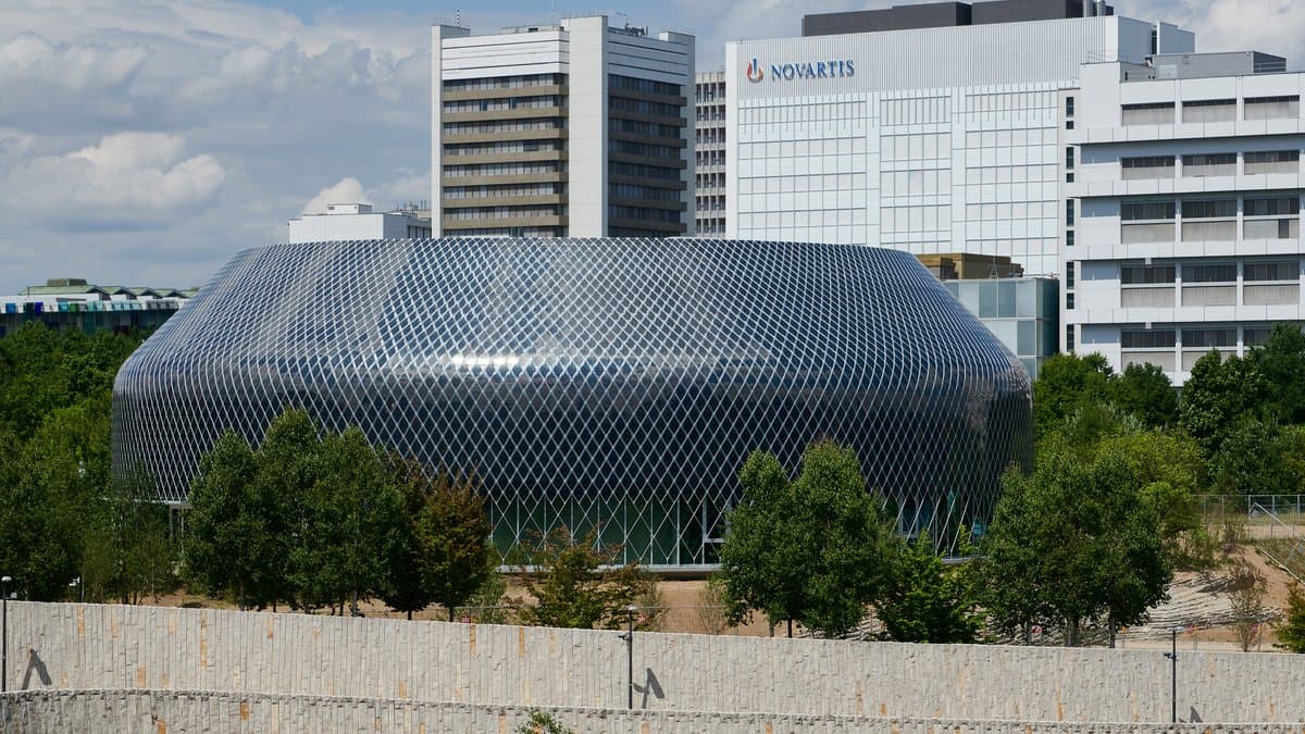 The silver, scale-clad Novartis Pavillon on the Novartis campus in Basel, Switzerland, with an office block carrying the Novartis name behind it.