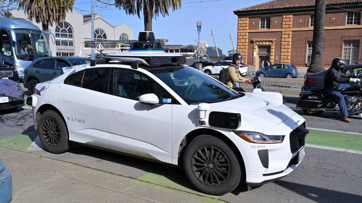 A white driverless Jaguar I-Pace with roof-mounted lidar and camera sensors moves along a San Francisco street beside parked cars, a motorcyclist and people on the pavement.