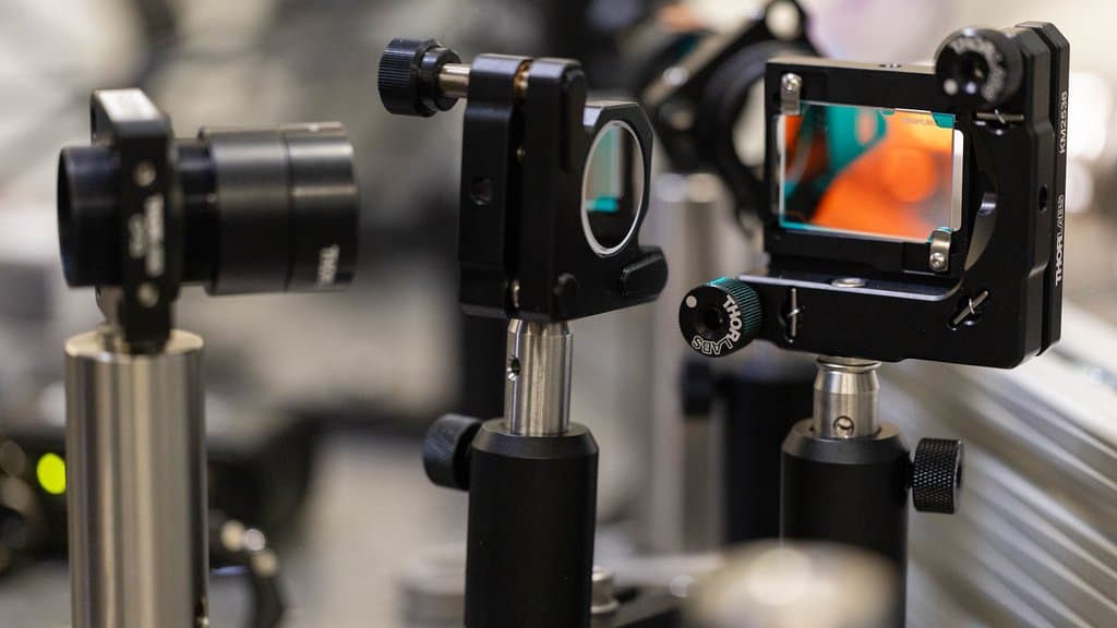 Close-up of black optical mounts, lenses and a dichroic filter standing on posts on an optical bench, part of a scanning nitrogen-vacancy diamond microscope.
