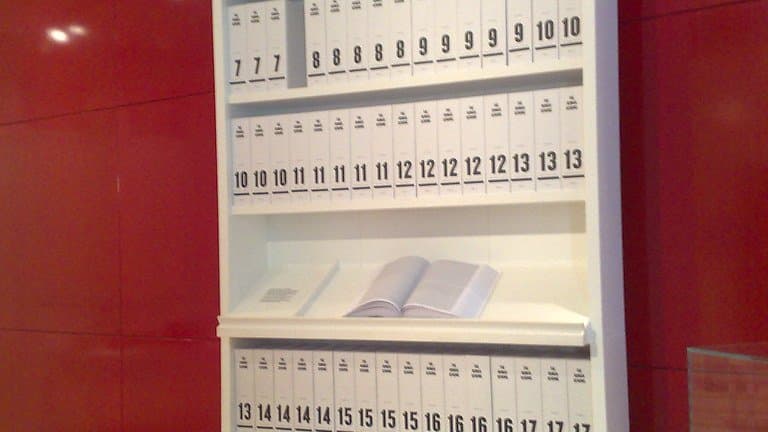 A tall white bookcase against a red wall holding rows of identical slim white volumes labelled with chromosome numbers, with one volume lying open on a middle shelf.