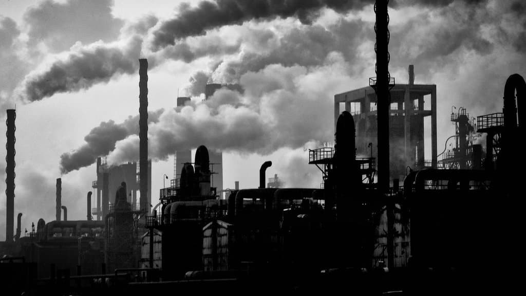 Black-and-white photograph of an industrial plant whose tall chimneys pour dense smoke plumes across the sky above silhouetted pipework and towers.