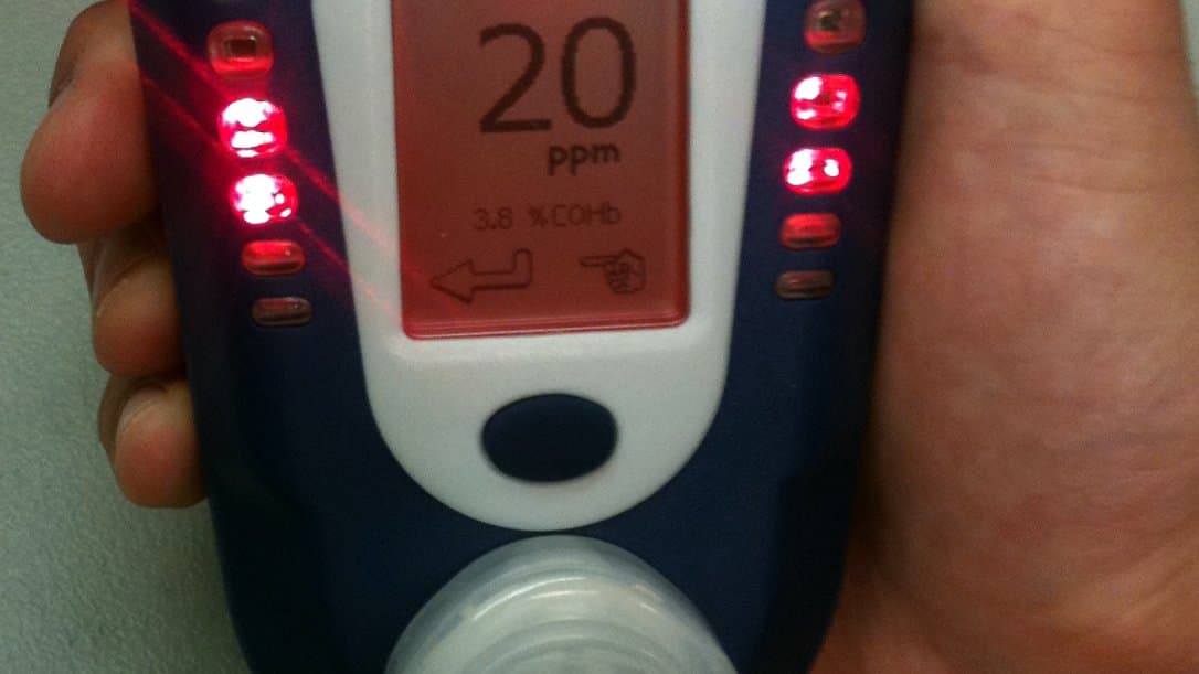 A hand holds a small breath carbon monoxide monitor whose screen reads 20 ppm and 3.8 percent COHb, with red indicator lights lit along both sides of the display.