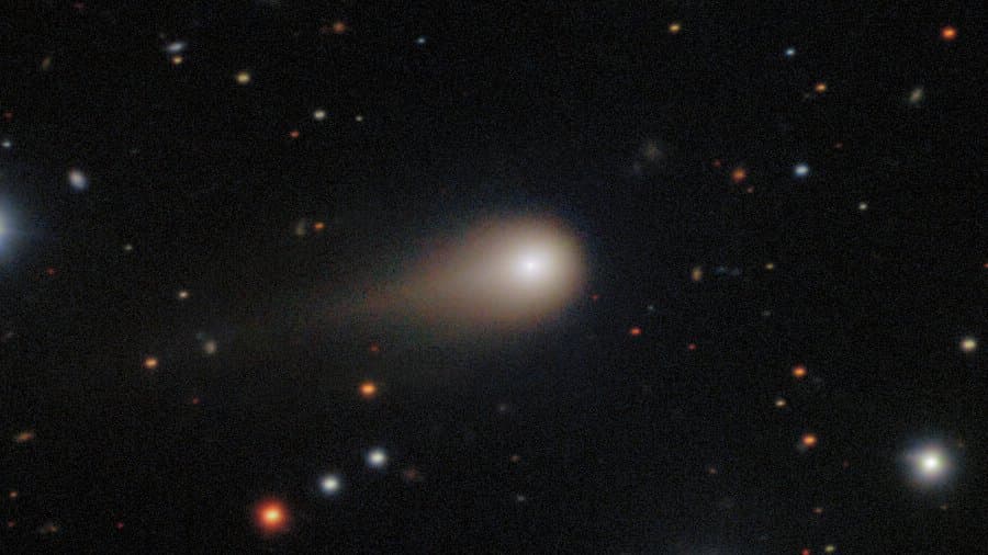 Colour telescope image of interstellar comet 3I/ATLAS: a bright condensed coma with a broad tail streaming to the left, set against a field of stars and small distant galaxies.
