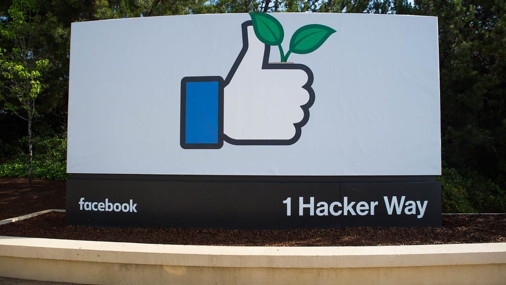 The thumbs-up entrance sign at 1 Hacker Way, Meta's headquarters campus in Menlo Park, California, with 'facebook' and '1 Hacker Way' lettering along its base.