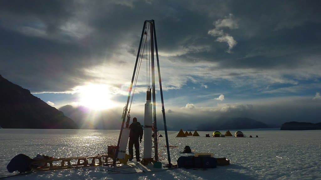 A tripod ice-core drill and a small field camp silhouetted on a snow-covered glacier surface, with the low sun behind them.