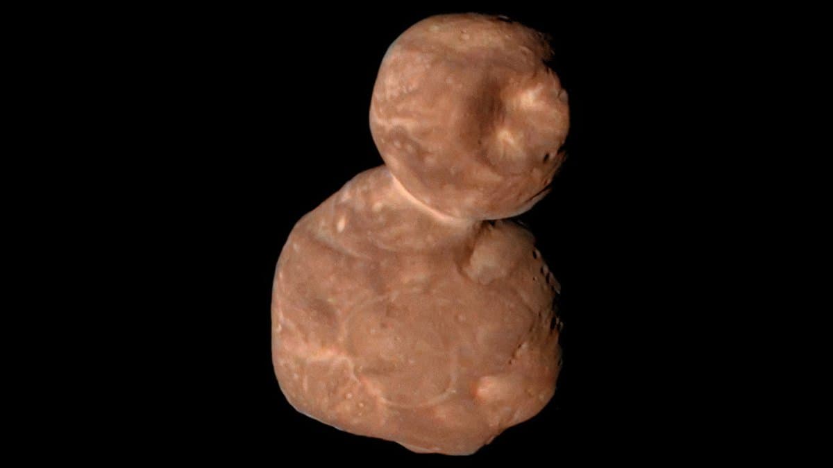 The two-lobed, reddish icy body Arrokoth against black space, a small round lobe joined to a larger flattened one, its surface mottled with bright patches and shallow pits.