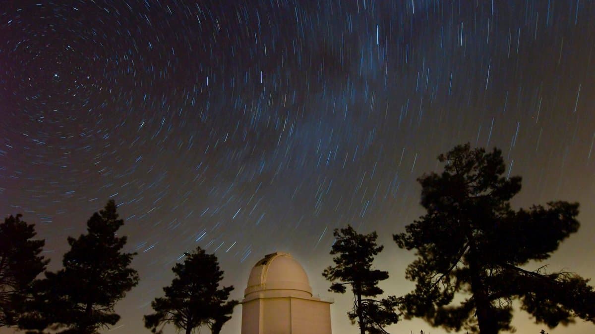 A white observatory dome on a pine covered ridge beneath a night sky filled with circular star trails.