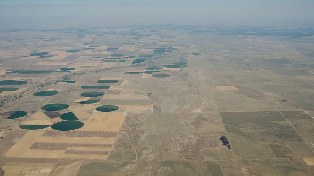 Aerial view of flat farmland stretching to a hazy horizon, with dark green circles of center-pivot irrigation scattered among pale, dry fields.