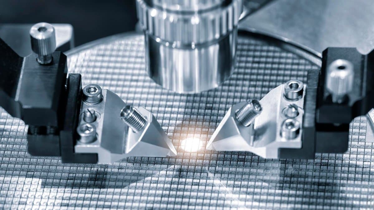 Metal tooling and an optical column positioned over a patterned silicon wafer, with a bright point of light where the tool meets the surface.