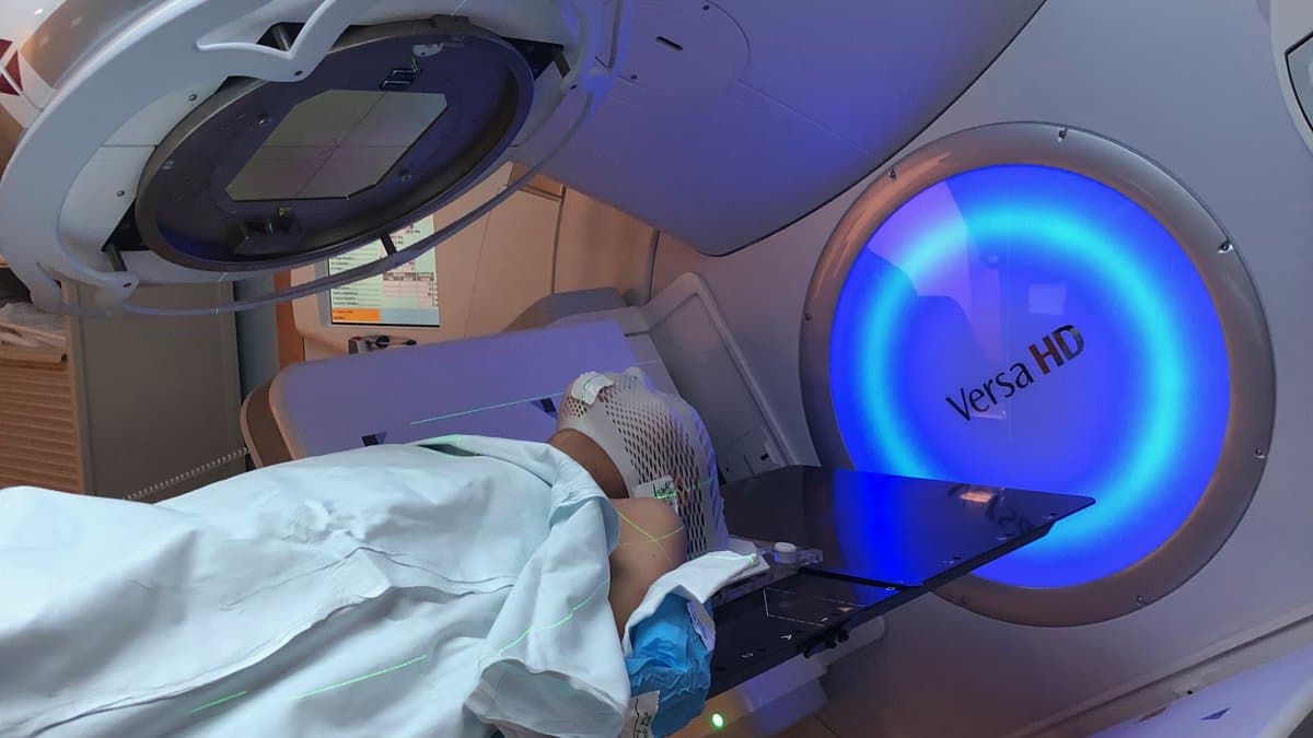 A patient lies on the treatment couch of a linear accelerator, with the machine's gantry and imaging panel positioned over them.