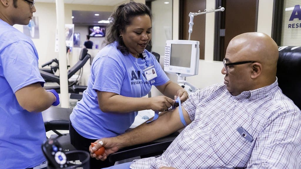 A nurse wearing an All of Us Research Program shirt draws a blood sample from a seated participant at an enrollment site while a second staff member assists.