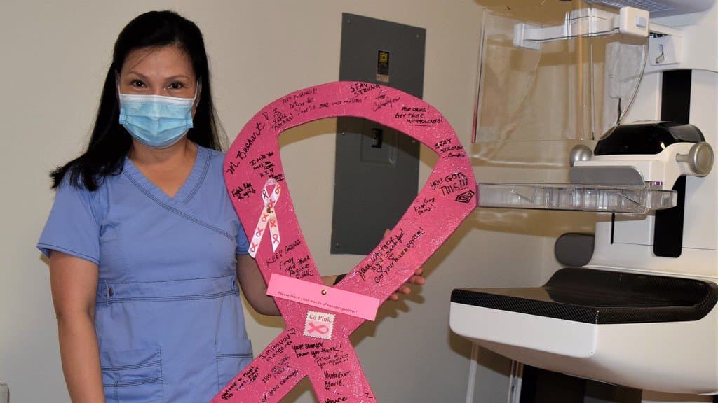 A technologist in scrubs and a face mask holds a large pink ribbon covered in handwritten signatures, standing beside a mammography machine.
