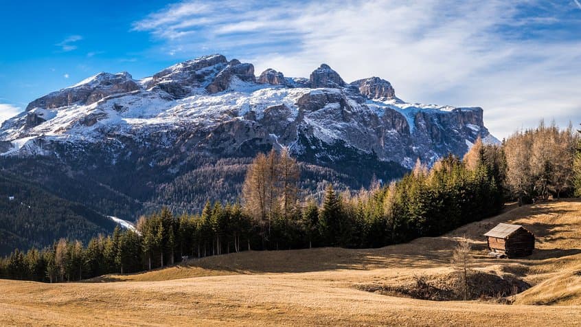 A wide view of a Dolomite massif with patchy snow on its upper cliffs, above a dry golden meadow, larch woods and a small wooden barn.
