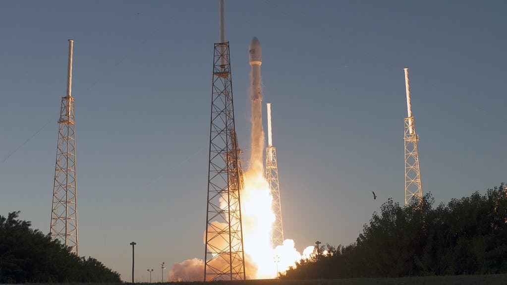 A SpaceX Falcon 9 rocket lifts off at dusk from its pad at Cape Canaveral, its exhaust plume glowing between the pad's tall lightning towers.