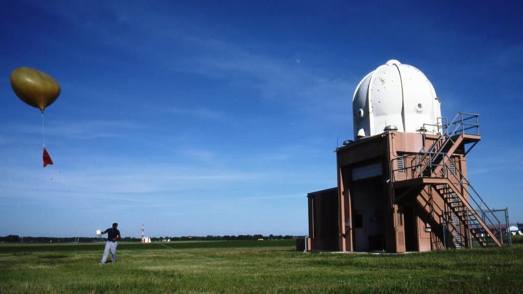 An observer stands on a grass field releasing a weather balloon that carries a small radiosonde package, with a domed weather radar tower on the right.