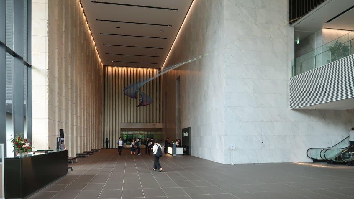 The tall, marble-lined lobby of a modern Tokyo office tower, with a reception desk at left and a few people walking toward the entrance.