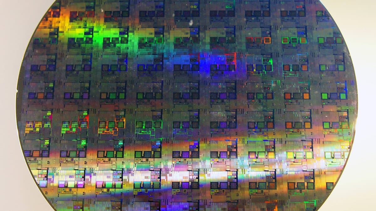 A circular silicon wafer covered in a grid of rectangular integrated circuit dies, diffracting light into green, blue and orange bands.
