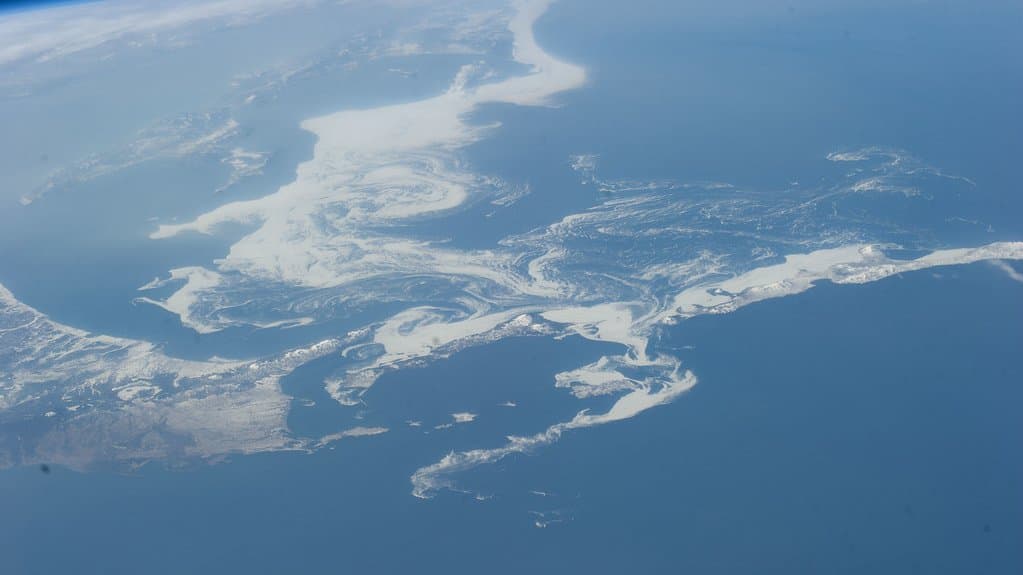 Photograph from the International Space Station showing pale swirls of drift ice spread across the dark blue Sea of Okhotsk, with snow-covered coastline and islands along its edges.