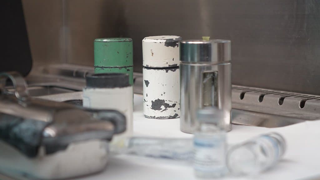 Lead shielding pots and small unlabeled glass vials on a white bench inside a stainless steel radiopharmacy hood.