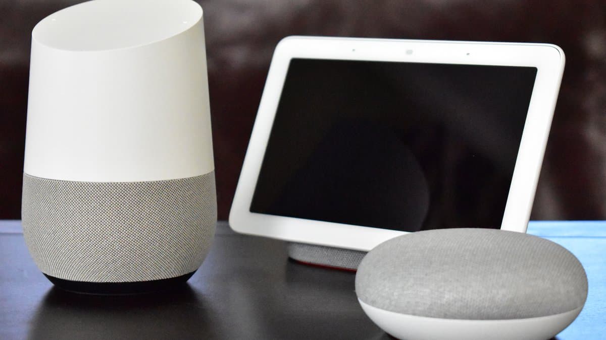 Three Google voice-assistant devices on a table: a cylindrical Home speaker, a Home Hub smart display with a dark screen, and a puck-shaped Home Mini.