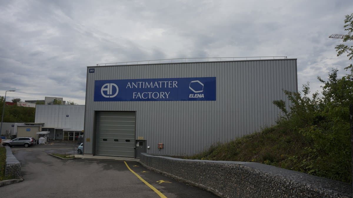 The CERN Antimatter Factory, a gray metal hall with a large blue sign reading ANTIMATTER FACTORY and ELENA above a roll-up vehicle door, with a paved access road running up to it.