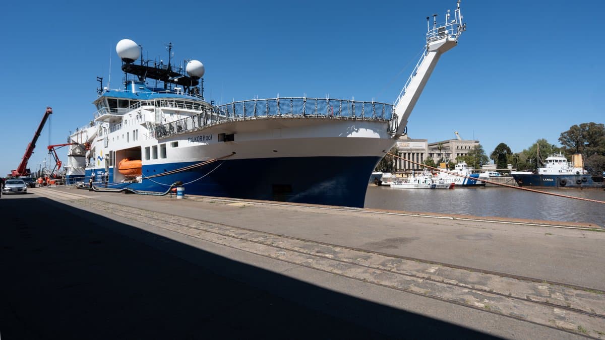 The research vessel Falkor (too) moored at a quay, its A-frame crane extended over the water under a clear sky.