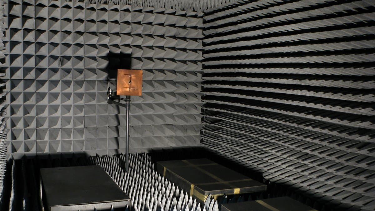 A small antenna on a stand inside a radio-frequency anechoic chamber, facing walls covered in gray pyramidal radio-absorbing foam.