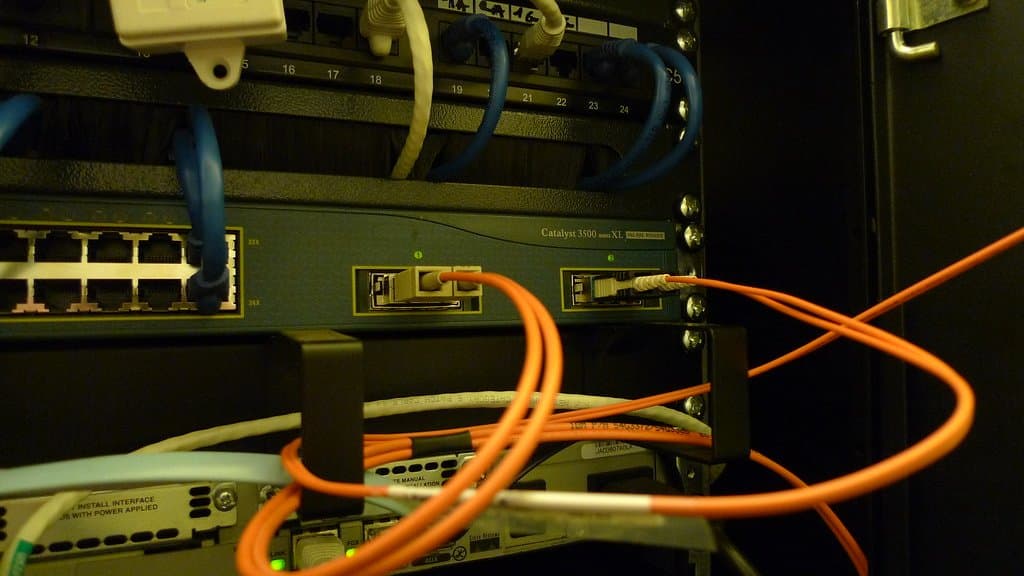 A rack-mounted Cisco Catalyst 3500 series switch with blue patch cables plugged into a panel above it and orange fiber cables looping across the front of the rack.