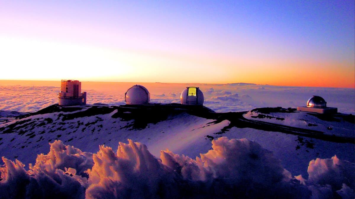 Telescope domes on a snow-dusted volcanic summit above a sea of clouds at sunset, with an orange and violet sky behind them.
