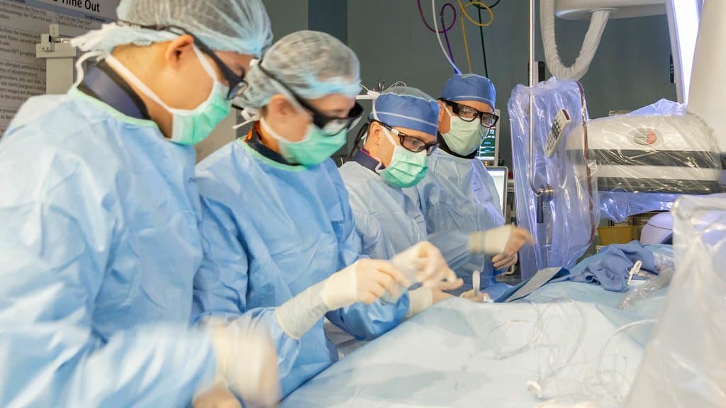 Four gowned and masked clinicians lean over a draped patient in a hospital catheterization laboratory, with an X-ray imaging arm and display monitors beside the table.