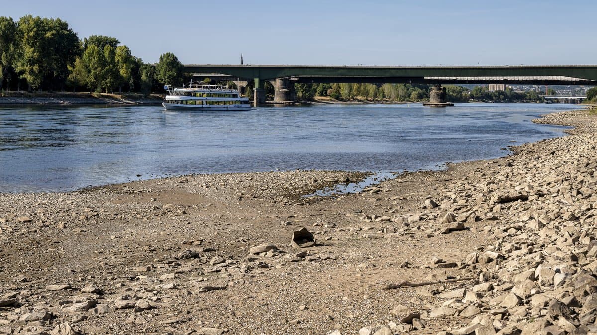 A broad bank of bare rock and gravel exposed along a river at low water, with a passenger vessel moving up the narrowed channel toward a road bridge.