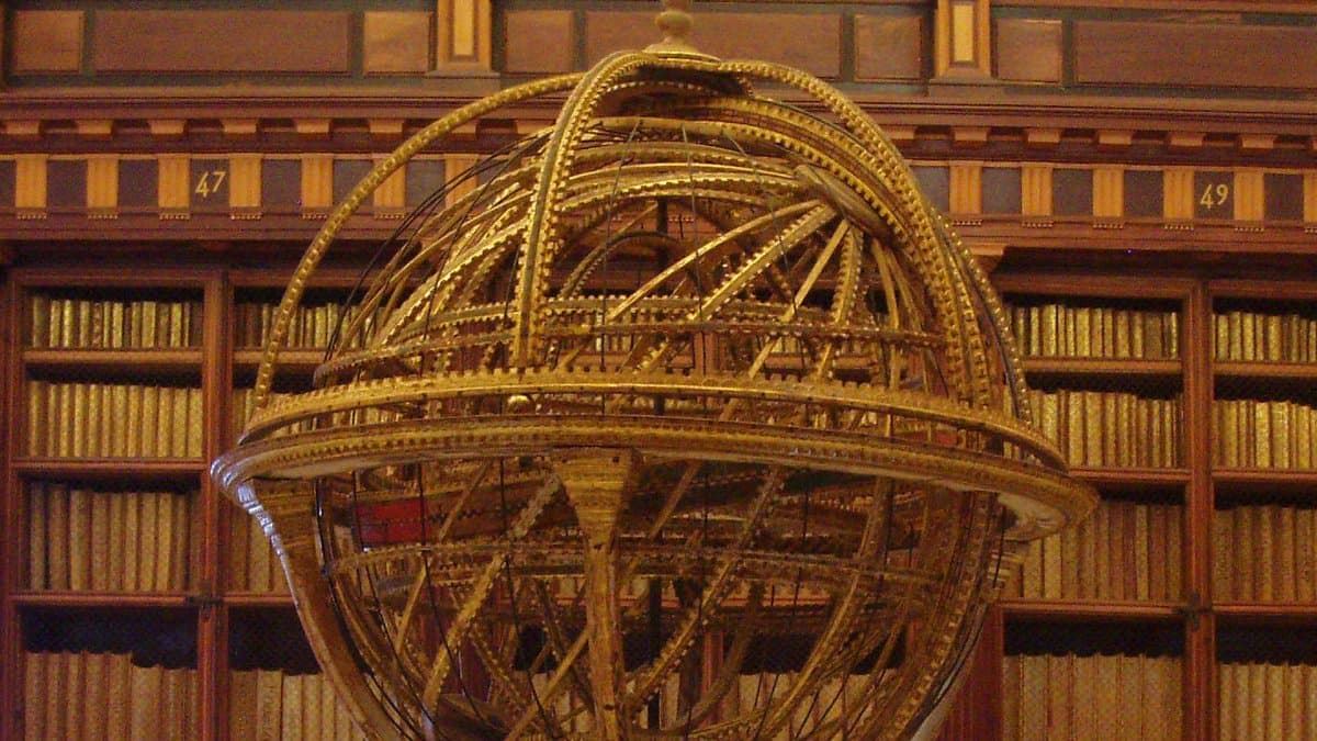 A large gilded armillary sphere of interlocking graduated rings standing in front of the wooden bookshelves of a historic library.