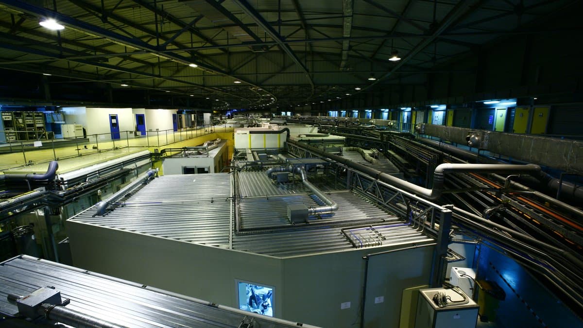 A long machine hall at a synchrotron facility, with the concrete-shielded storage ring running down the center and cable trays overhead.