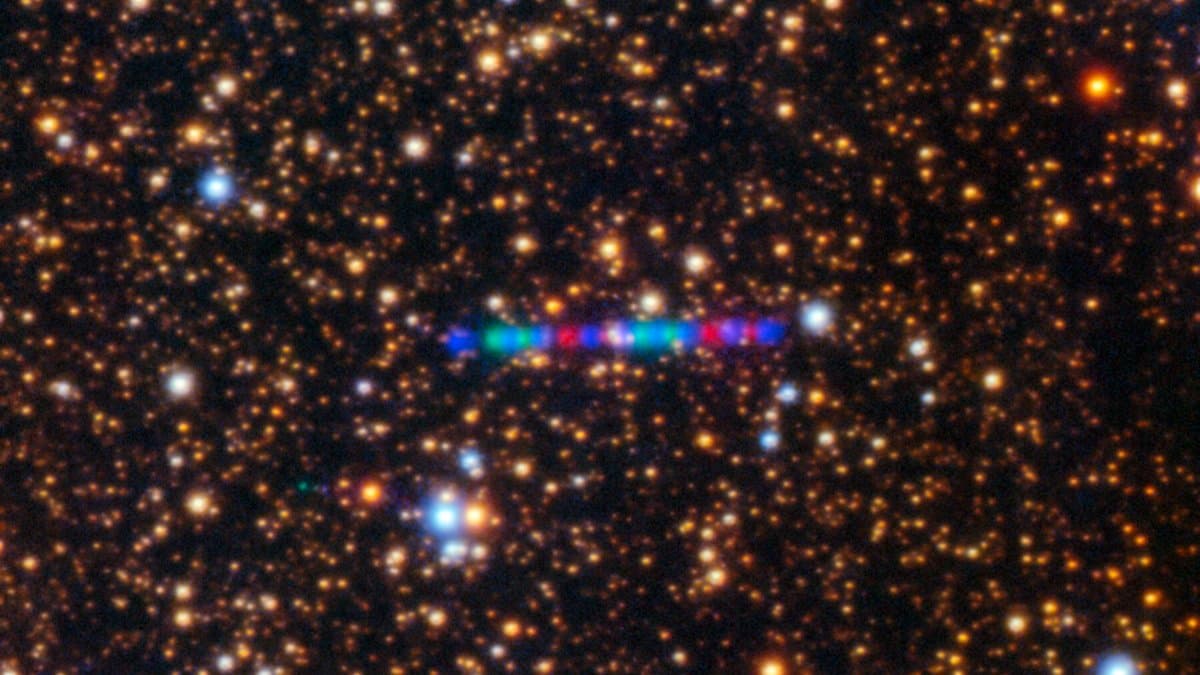 A dense field of orange and blue stars with a short, multicolored dashed streak near the center, which is the interstellar comet 3I/ATLAS.