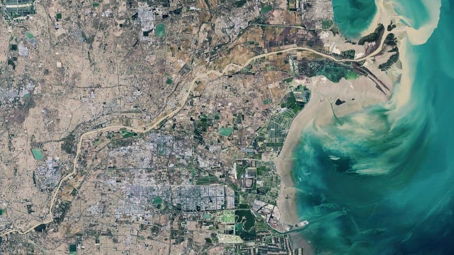 Satellite view of a low-lying delta coast, where a winding river channel crosses a flat, densely developed plain and sediment-laden turquoise water spreads over shallow tidal flats.